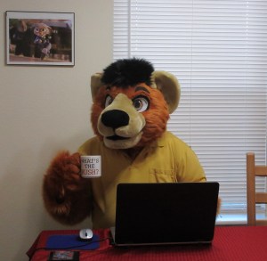 Person in a bear costume sitting at a dinner table behind a laptop. He is wearing a yellow polo shirt and holding a coffee mug with the words "What's the Rush?" on it. 